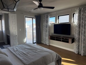 Ein Schlafzimmer mit Bett, Deckenventilator, an der Wand montiertem Fernseher und einer Glastür zum Balkon.