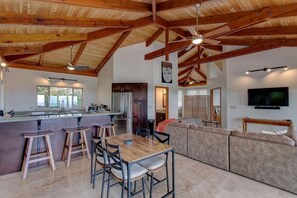 A spacious living area with a kitchen, wooden ceiling beams, a dining table with stools, a large sofa, and a flat-screen TV. - Lovely 2-bedroom house in Pāhoa perfect for a relaxing getaway (Pāhoa)