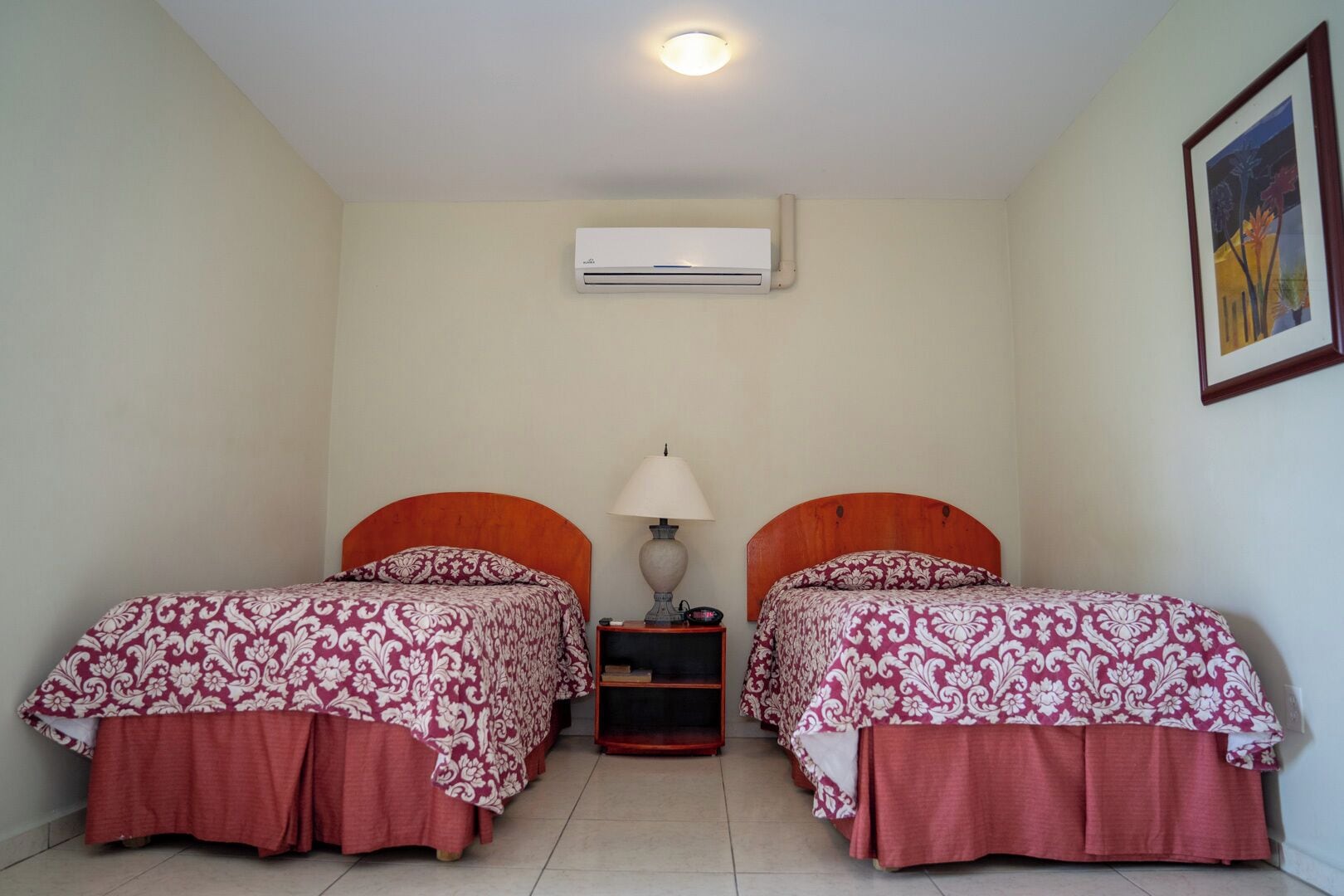 A room with two beds, each with a wooden headboard and a patterned bedspread. There is a nightstand with a lamp between the beds, and an air conditioning unit mounted on the wall.