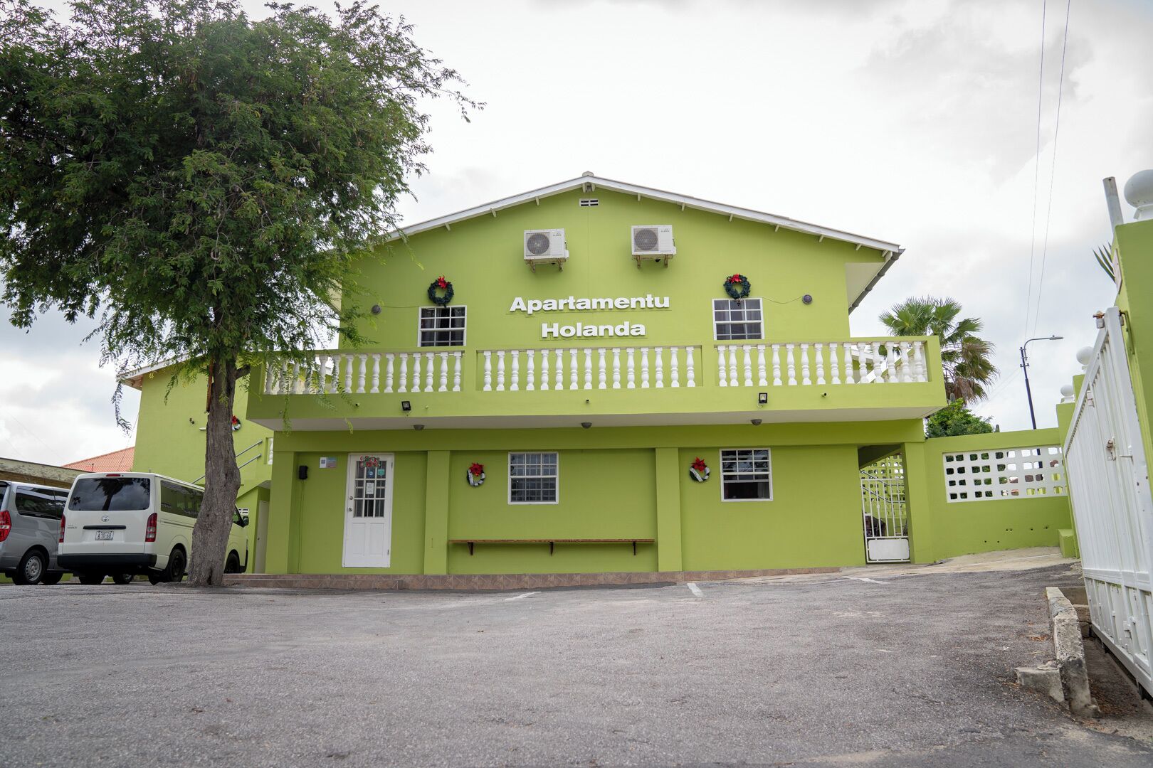 A two-story building with a green facade, labeled "Apartamento Holanda." It features a balcony with white railings, multiple windows, and air conditioning units.