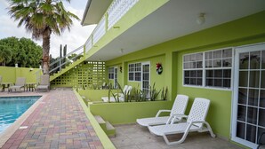 A poolside area with a tiled walkway, white lounge chairs, and a palm tree. - Apartamentu Holanda (Willemstad)