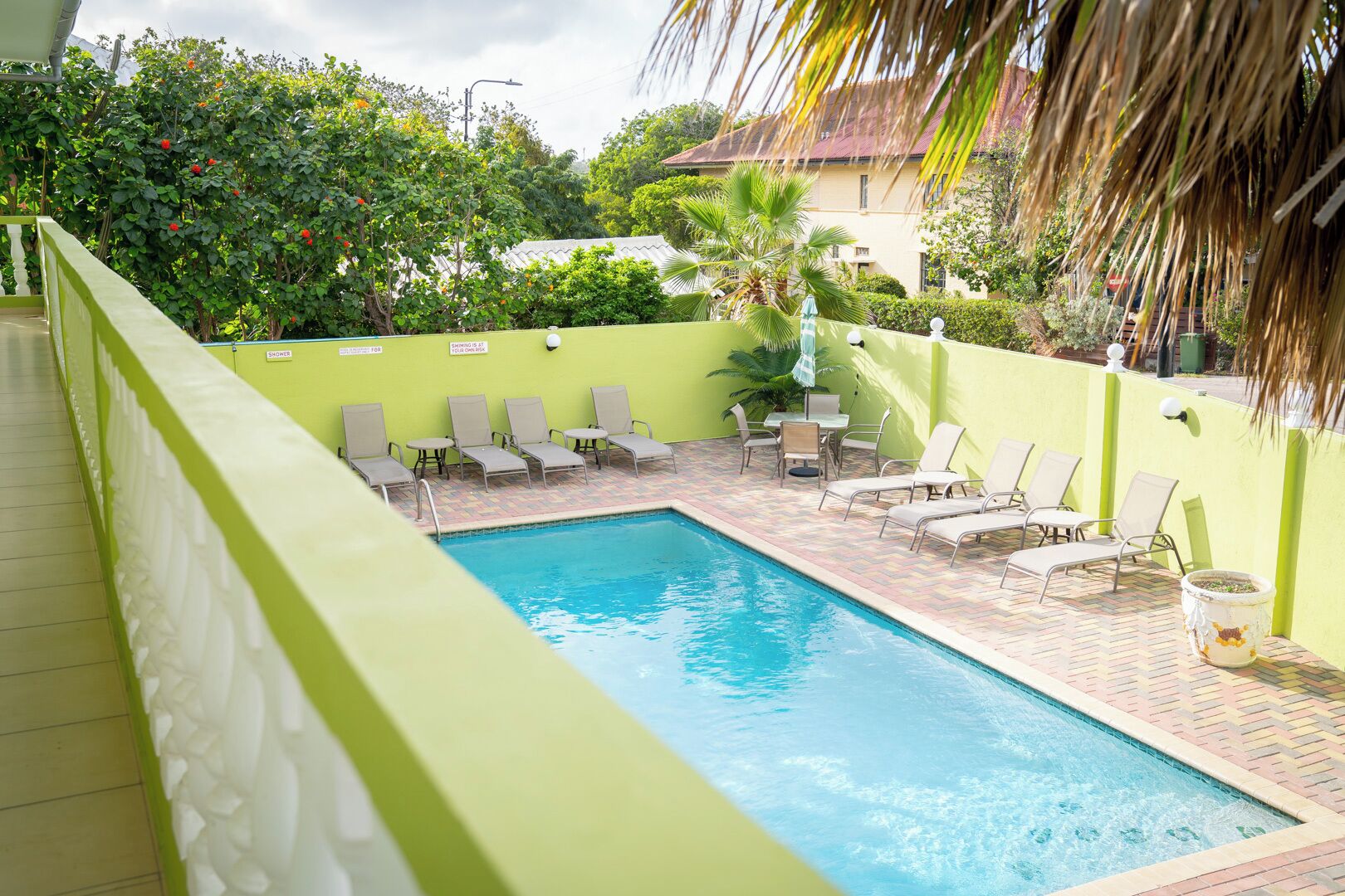 A swimming pool with lounge chairs and a table, surrounded by greenery and a building in the background.