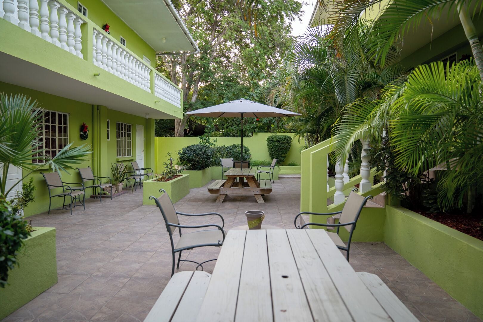 A courtyard with a wooden table and chairs, surrounded by greenery and a green building with white railings.