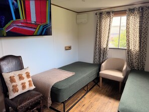 A room with a bed, a chair, a sofa, a wall-mounted air conditioner, a window with patterned curtains, and a painting.