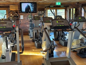 A gym with multiple cardio machines, a TV screen, and a brick wall.
