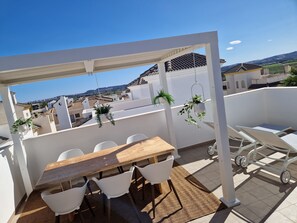 Eine Dachterrasse mit Holztisch und weißen Stühlen, umgeben von hängenden Pflanzen und einer weißen Pergola.