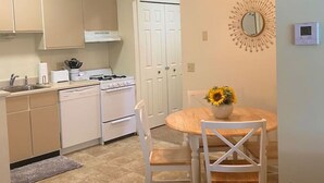 Cocina compacta con armarios blancos, estufa y ventilador de techo. Cuenta con una zona de comedor con mesa y sillas, además de un espejo decorativo en la pared.