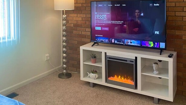 Una sala de estar con pared de ladrillo, un televisor de pantalla plana sobre un pedestal blanco, una lámpara de pie y una chimenea.