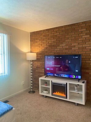 A living room with a brick wall, a flat-screen TV on a white stand, a floor lamp, and a fireplace. - Sunlit 1BR Near Pro Football HOF & Cak Airport (North Canton)