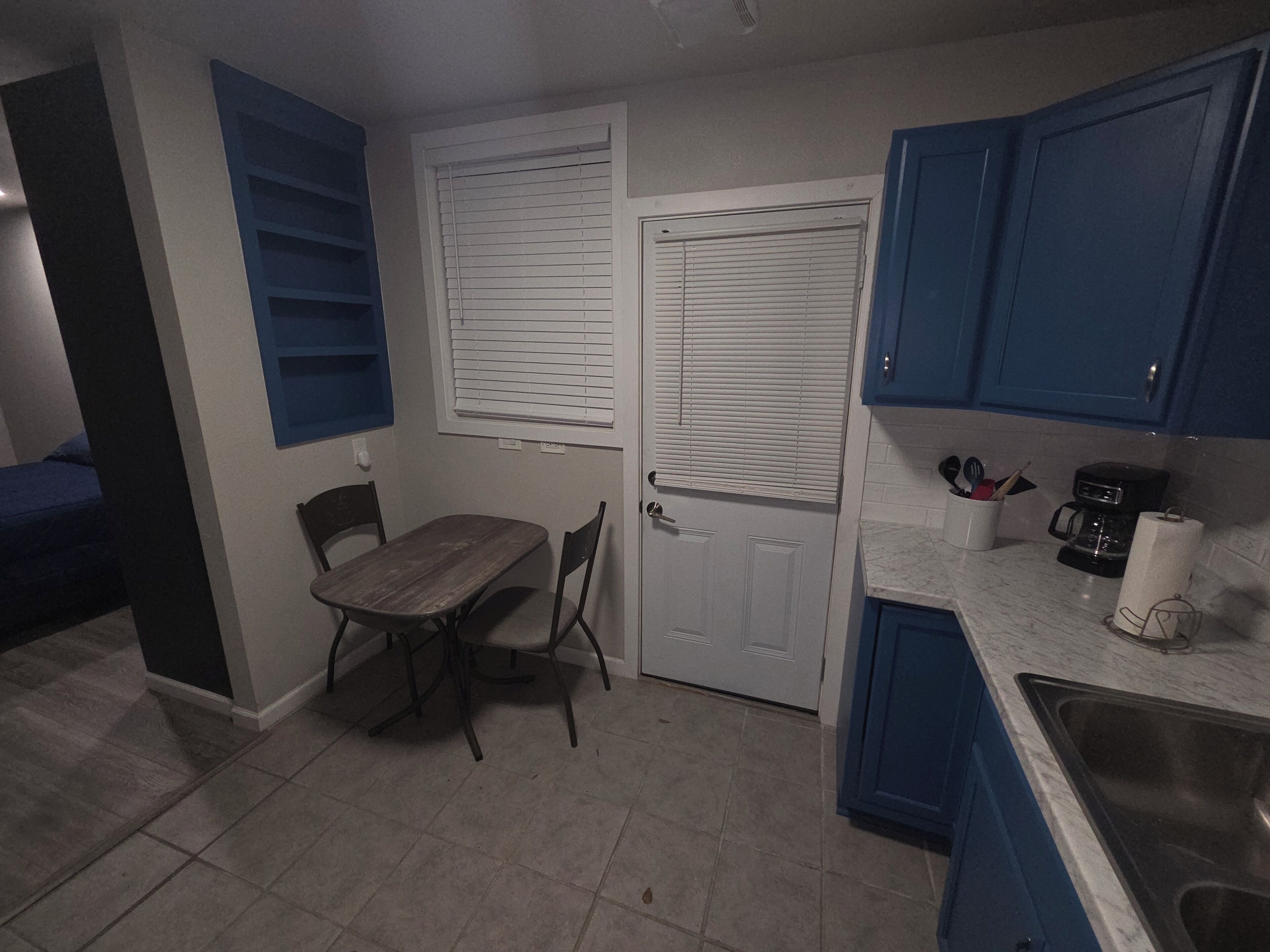 Une petite cuisine avec des placards bleus, une table en bois et une porte blanche avec des stores.