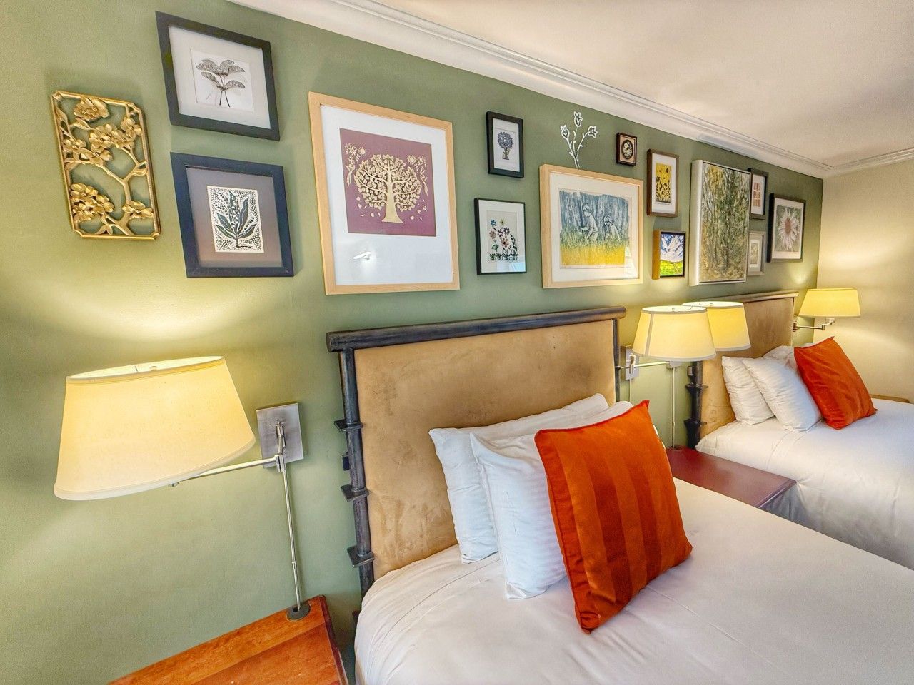 A hotel room with two beds, a headboard, wall-mounted lamps, and framed artwork on the wall.