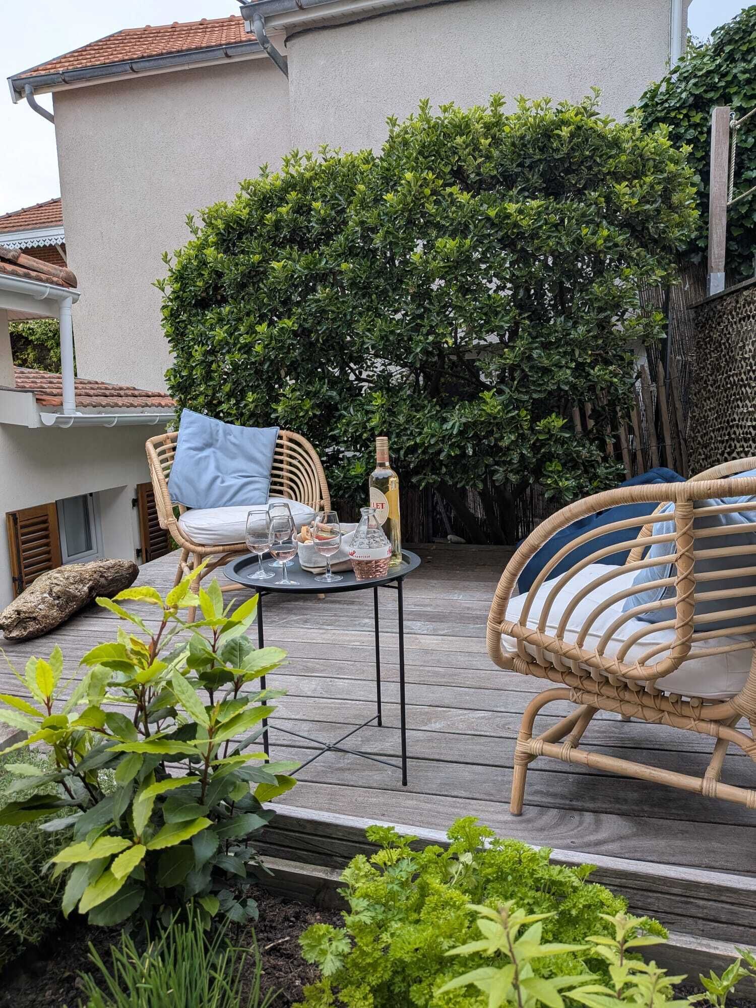 Eine Terrasse mit Korbmöbeln, einem Tisch mit Gläsern und einer Flasche, und ein üppiger, grüner Heckenstreifen im Hintergrund.