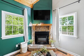 A room with a stone fireplace, a flat-screen TV mounted above it, and two windows with white frames. - Seconds from Helen Main St! Hot Tub - 4 bedrms - Sleeps 9 (Helen)