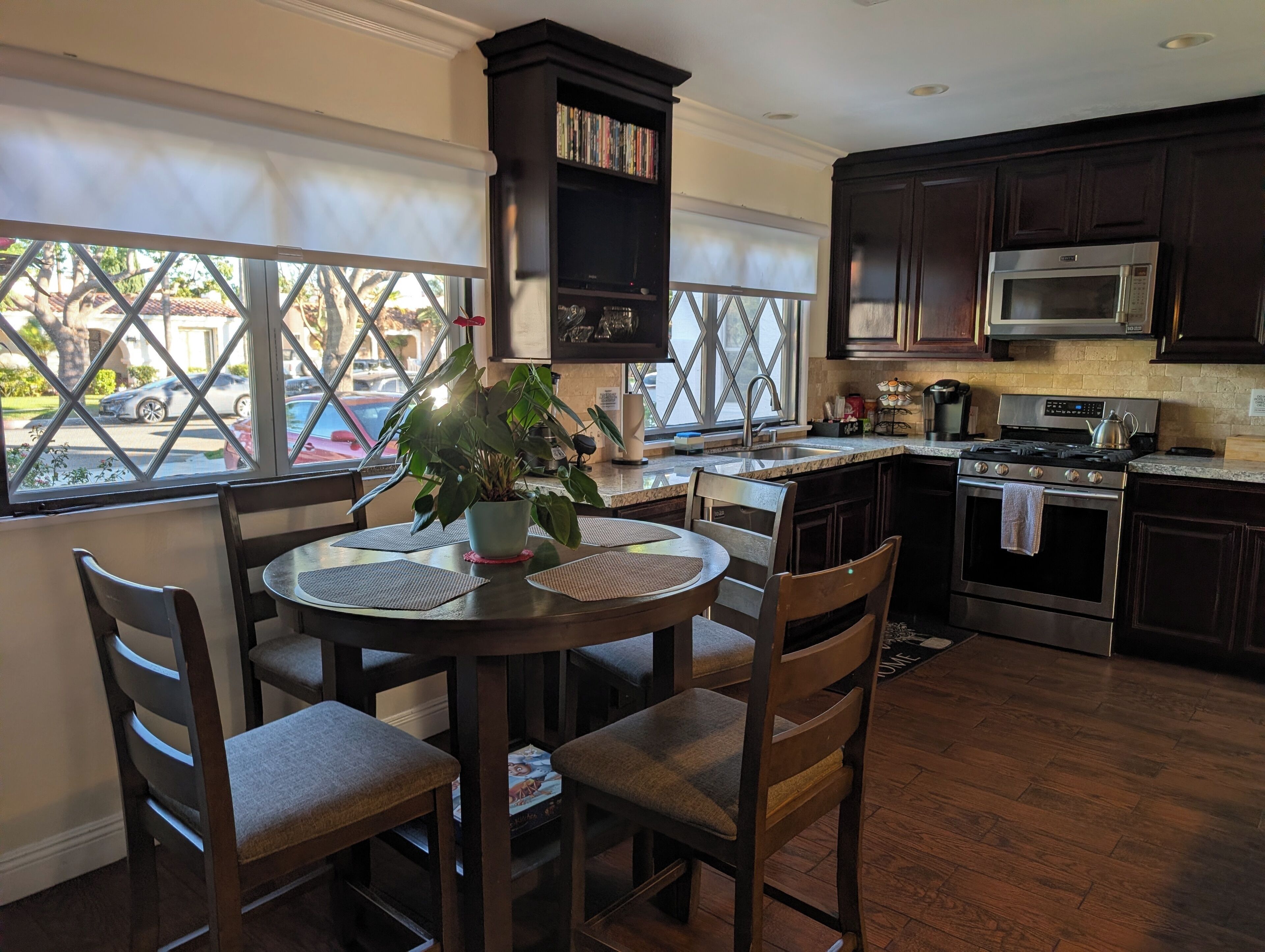 A kitchen with a dining area, featuring a round table with chairs, a potted plant, and a view through large windows.