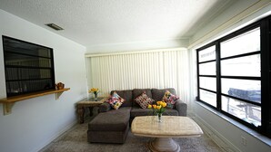Una sala de estar con un sofá modular, una mesa de centro redonda, una ventana con persianas, un estante y una mesita con un florero.