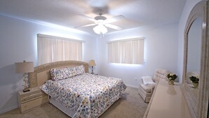 Un dormitorio con cama, mesita de noche, cómoda, espejo, ventilador de techo y dos ventanas con persianas.