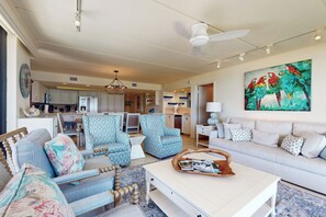 Ein geräumiges Wohnzimmer mit einem weißen Sofa, gemusterten Sesseln und einem Couchtisch mit Zeitschriften. Im Hintergrund ist eine Küche zu sehen, und ein farbenfrohes Gemälde ziert die Wand.