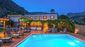Piscina con sillas de descanso y zona de bar, rodeada de vegetación y con montañas al fondo.