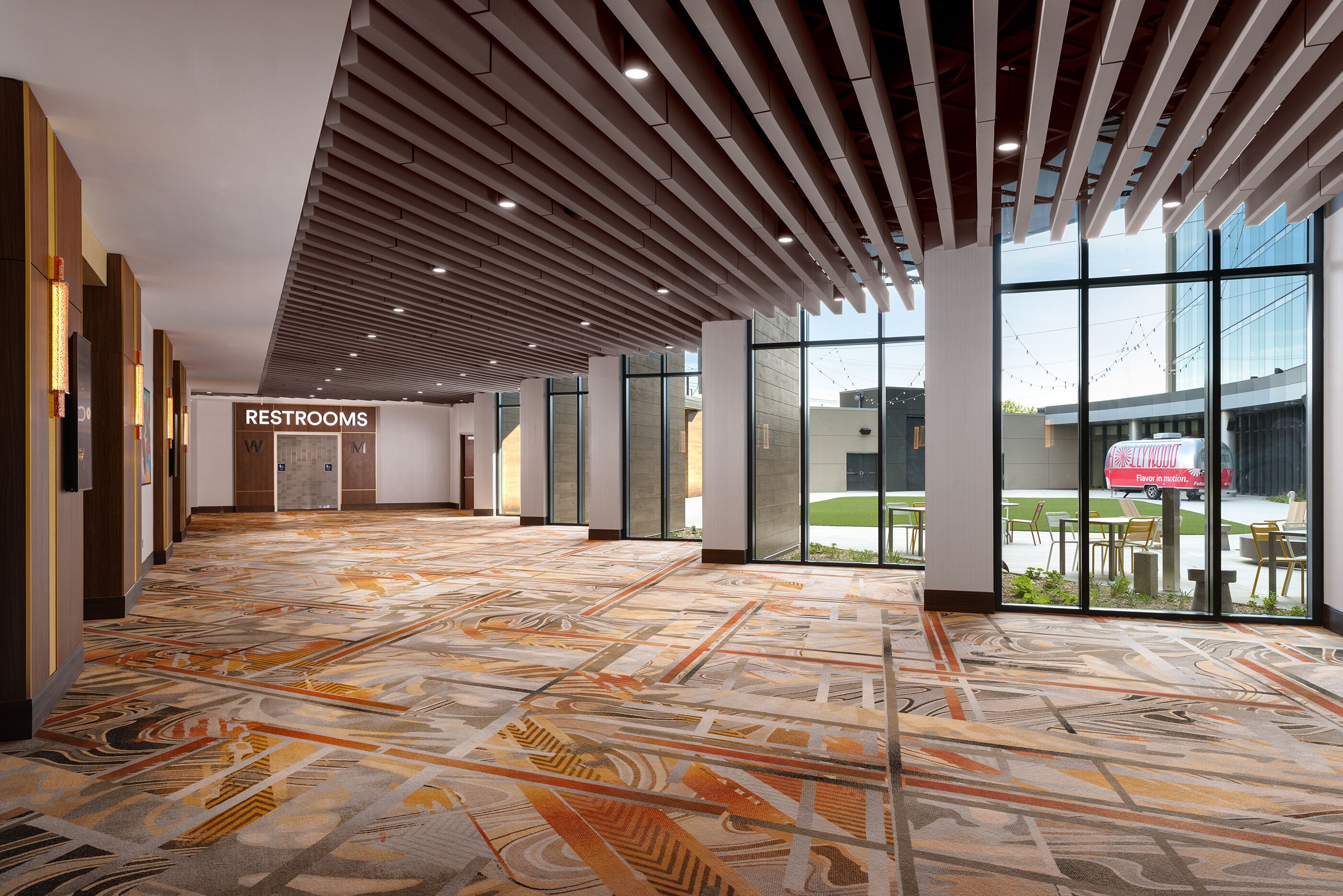 A spacious corridor with a patterned carpet, a sign indicating restrooms, and large windows offering views of the outside.