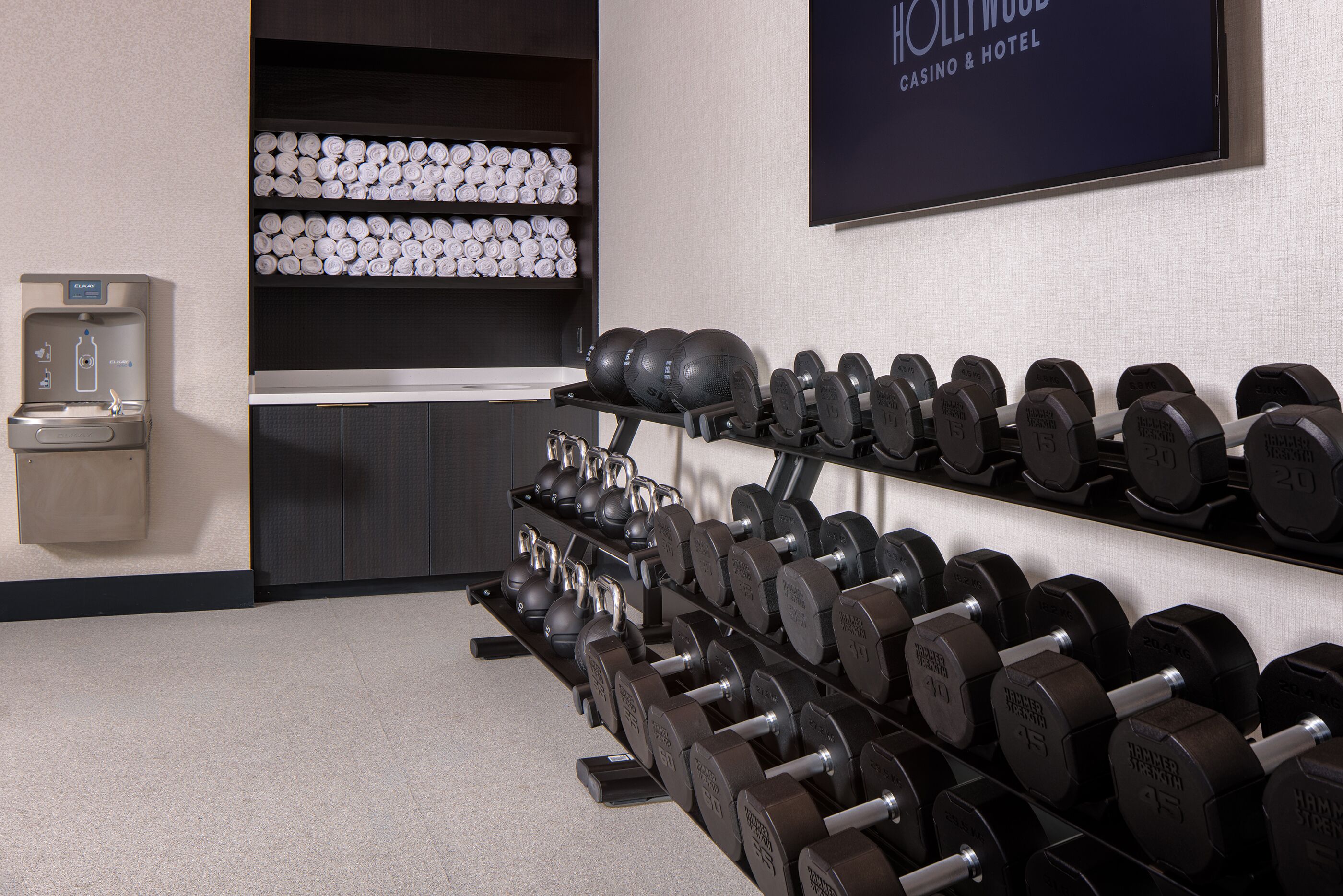 Un gimnasio con filas de mancuernas, un dispensador de agua y un televisor empotrado en la pared mostrando 'HOLLYWOOD CASINO & HOTEL'.