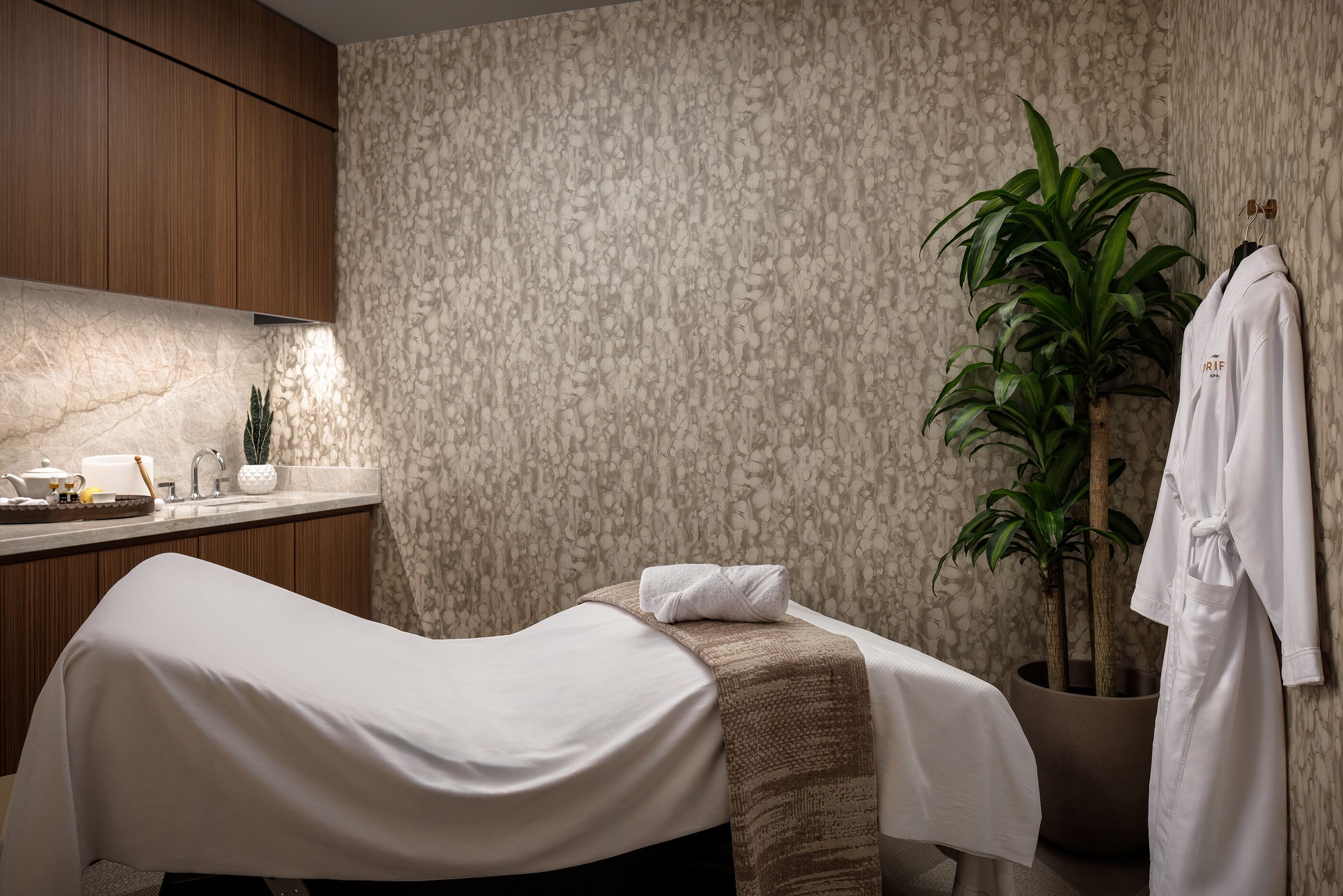 A spa room with a massage table, a towel, a robe, a plant, and a sink with toiletries.