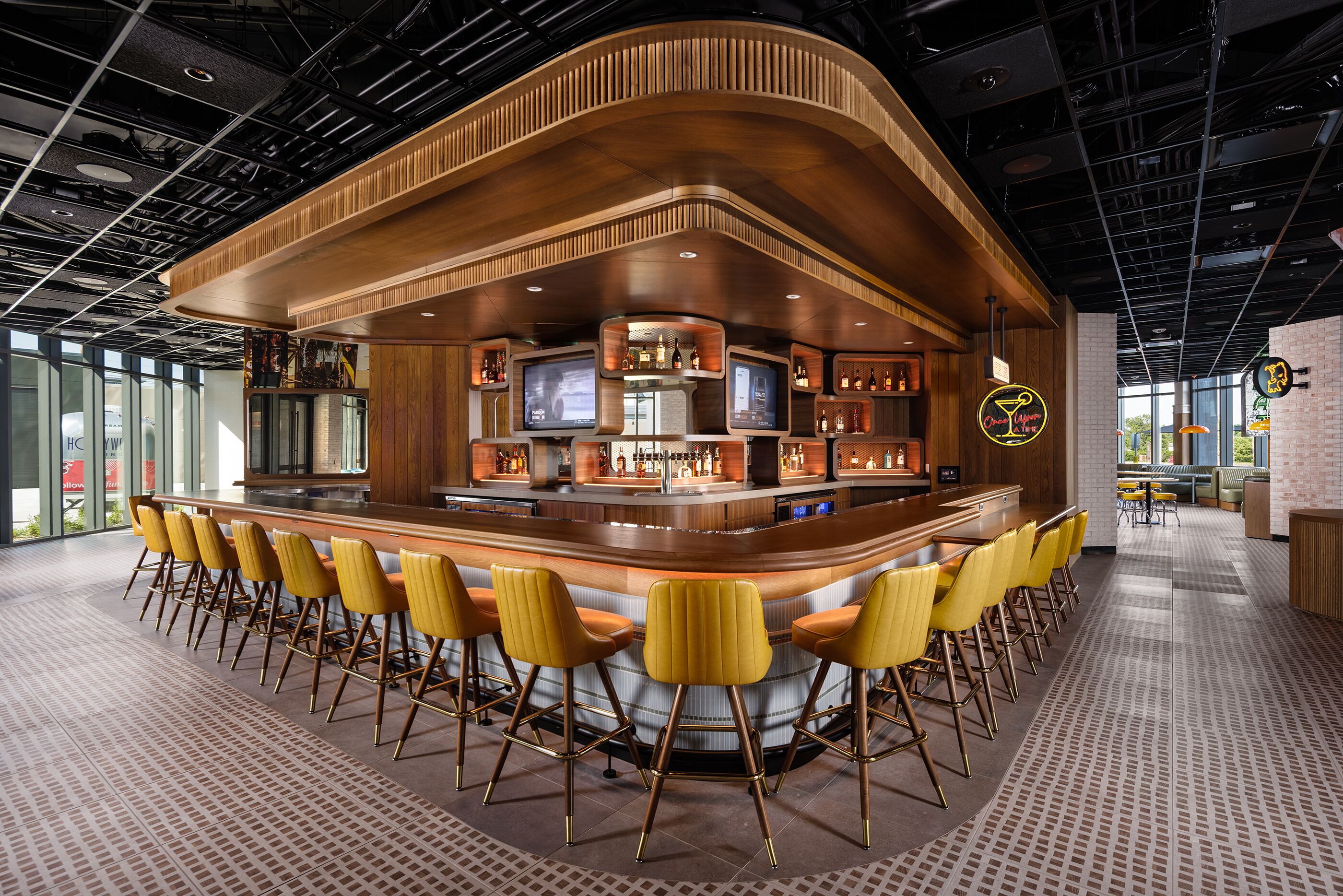 Uma área de bar com balcão curvo, várias telas e disposição de assentos com cadeiras amarelas.