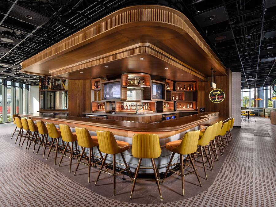 Uma área de bar com balcão curvo, várias telas e disposição de assentos com cadeiras amarelas.