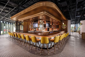 Zona de bar con barra curva, varias pantallas y disposición de asientos con sillas amarillas.