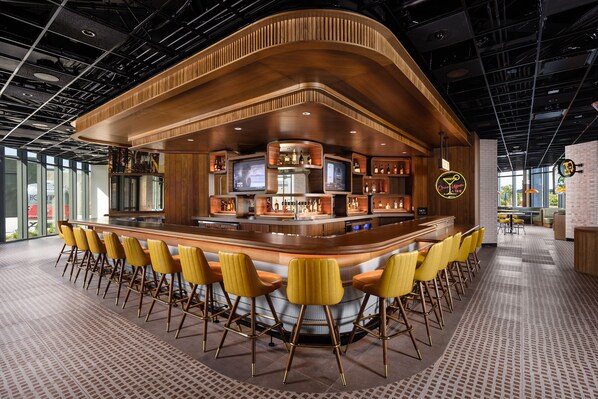 A bar area with a curved counter, multiple screens, and a seating arrangement with yellow chairs.