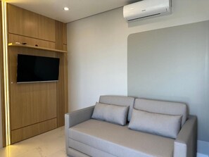 A modern living room with a flat-screen TV mounted on a wooden wall, a grey sofa with cushions, and an air conditioning unit. - Mirante do Oceano guesthouse, 70 meters from the beach, with a heated pool. (Bombinhas)