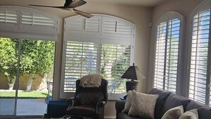 A living room with a ceiling fan, a sofa, a chair, and large windows with shutters. - Desert Escape in Indian Palms | Golf Cart Included | Near Coachella & Tennis Gardens #051797 (Indio)