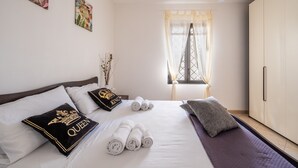 Ein Hotelzimmer mit Bett, Kissen, aufgerollten Handtüchern, einem Fenster mit Vorhängen, einem Schrank und Wandbildern.