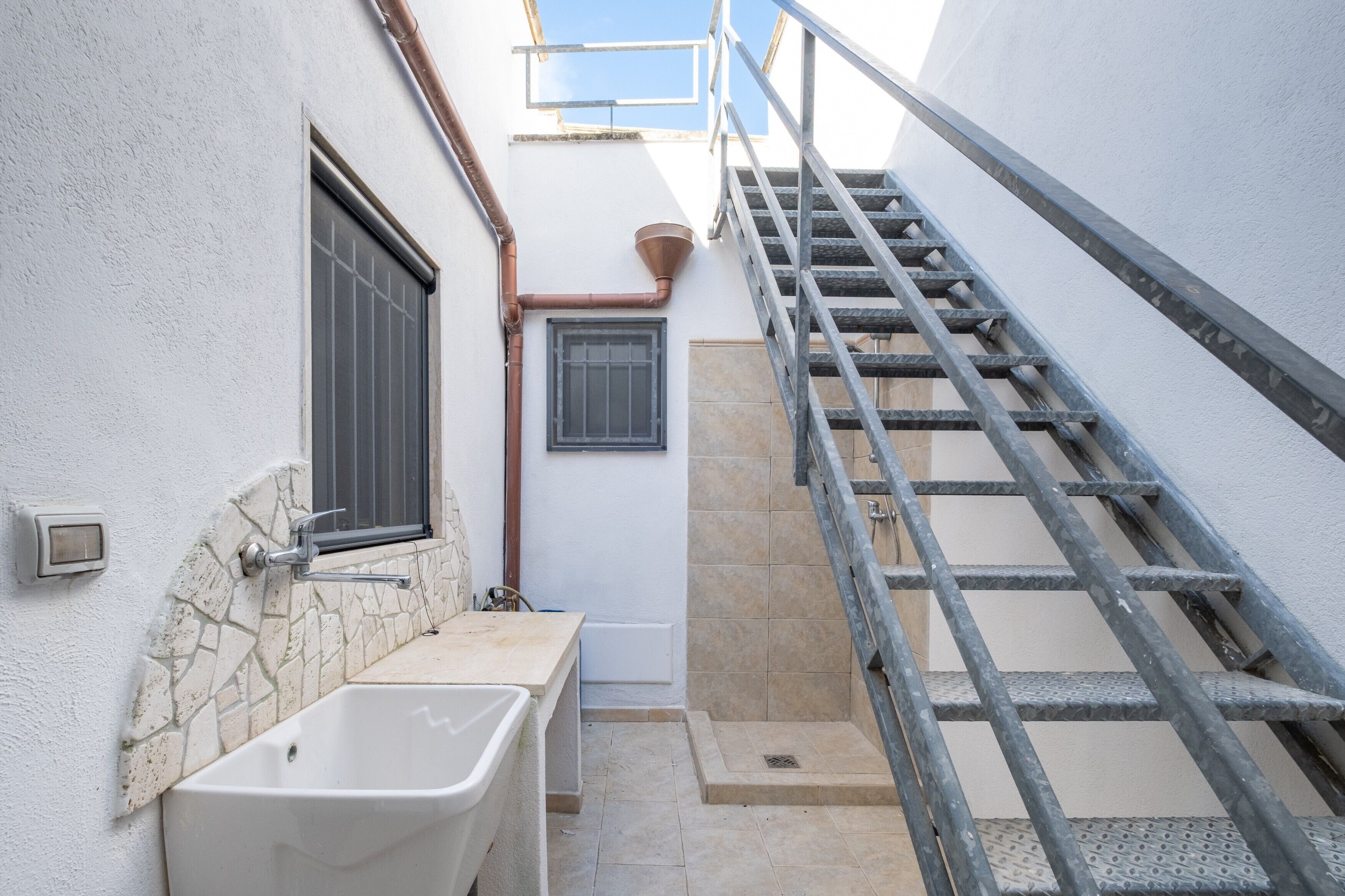Una escalera estrecha al aire libre que sube a un nivel superior, una bañera blanca y una ventana con barrotes.