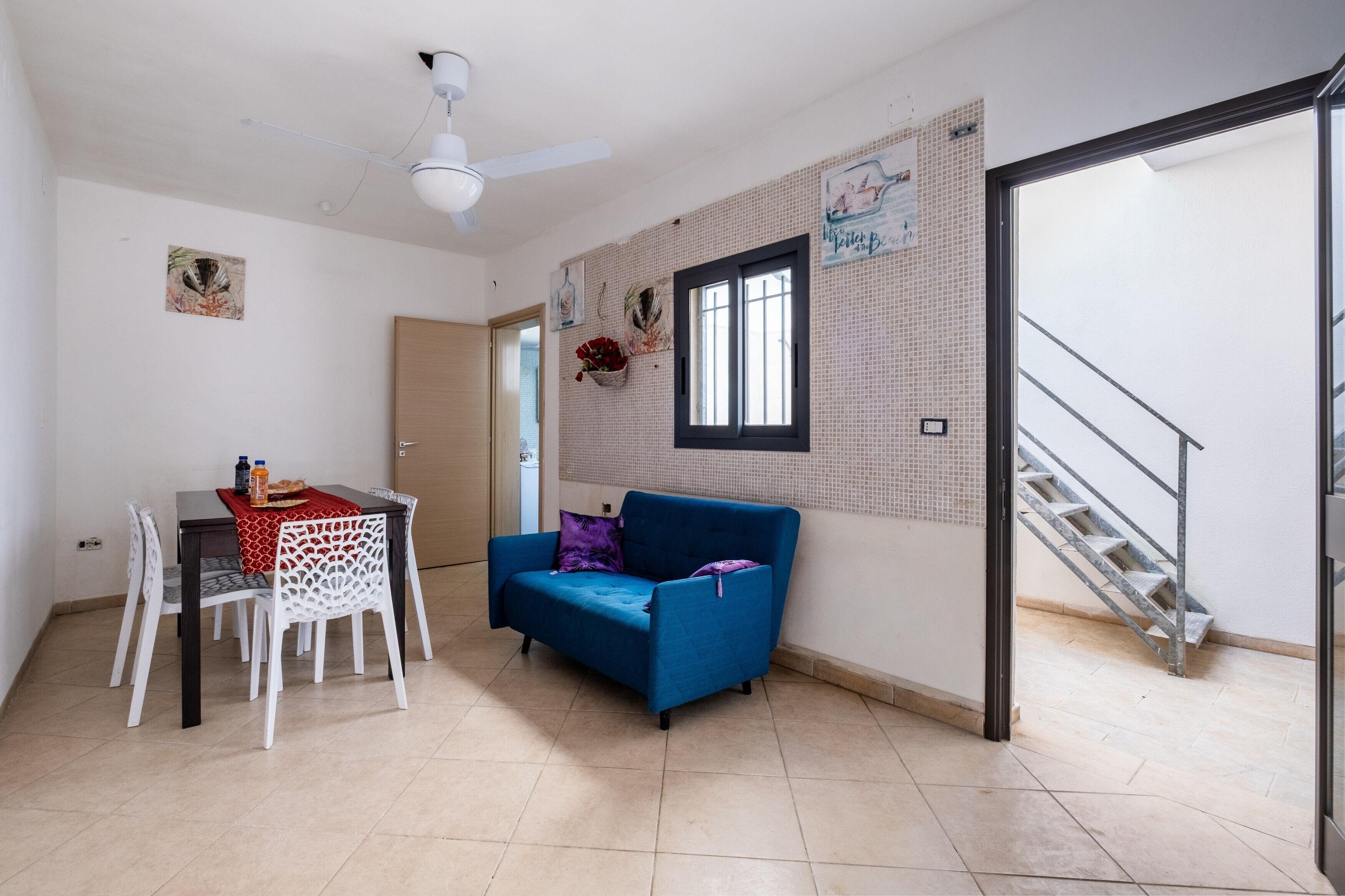 Ein Zimmer mit Esstisch, Stühlen, einem blauen Sofa, einem Deckenventilator, einem Fenster, einem Wandkorb mit Blumen und einer Treppe.