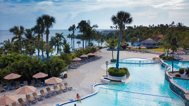 Piscina de resort com espreguiçadeiras e guarda-sóis, cercada por palmeiras e vista para o oceano.