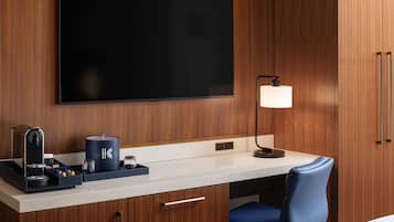 Habitación de hotel con televisor de pantalla plana, escritorio con cafetera, lámpara y silla azul.