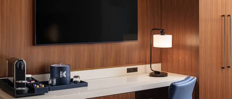 A hotel room with a flat-screen TV, a desk with a coffee maker, a lamp, and a blue chair.