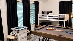 Un lit superposé, une table de billard et un porte-queue de billard dans une pièce avec des rideaux noirs et une fenêtre équipée de stores.