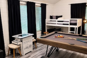 Een stapelbed, een pooltafel en een poolkeu-rek in een kamer met zwarte gordijnen en een raam met jaloezieën.