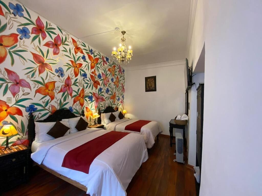 A hotel room with two beds, a floral wallpaper, a chandelier, and a bedside table with a lamp.