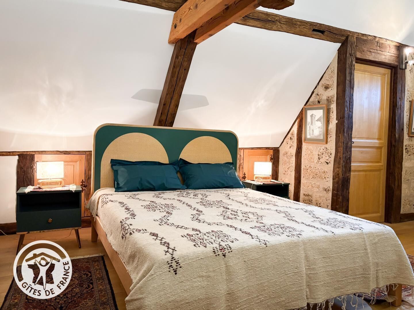 Ein Schlafzimmer mit hohen Decken, freiliegenden Holzbalken, einem Bett mit gemustertem Überwurf, zwei Nachttischen mit Lampen und einer Tür mit Spiegel.