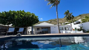 Una piscina con agua cristalina, un edificio blanco con un porche cubierto y una palmera.