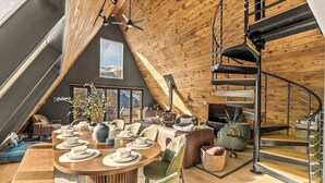 A spacious loft-style living area with a dining table set for four, a cozy seating area, a fireplace, and a spiral staircase. - Ultimate A-frame, 12 mi to Breck, Mtn View/Sauna/HT (Fairplay)