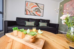 Un salon moderne avec un canapé noir, des coussins décoratifs, une table basse en bois agrémentée de plantes, un verre de jus d’orange et un tableau accroché au mur.
