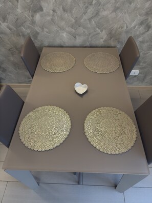 Una mesa rectangular con cuatro sillas, cada una con un cojín marrón. La mesa tiene un objeto blanco en forma de corazón en el centro y cuatro posavasos redondos y texturizados.