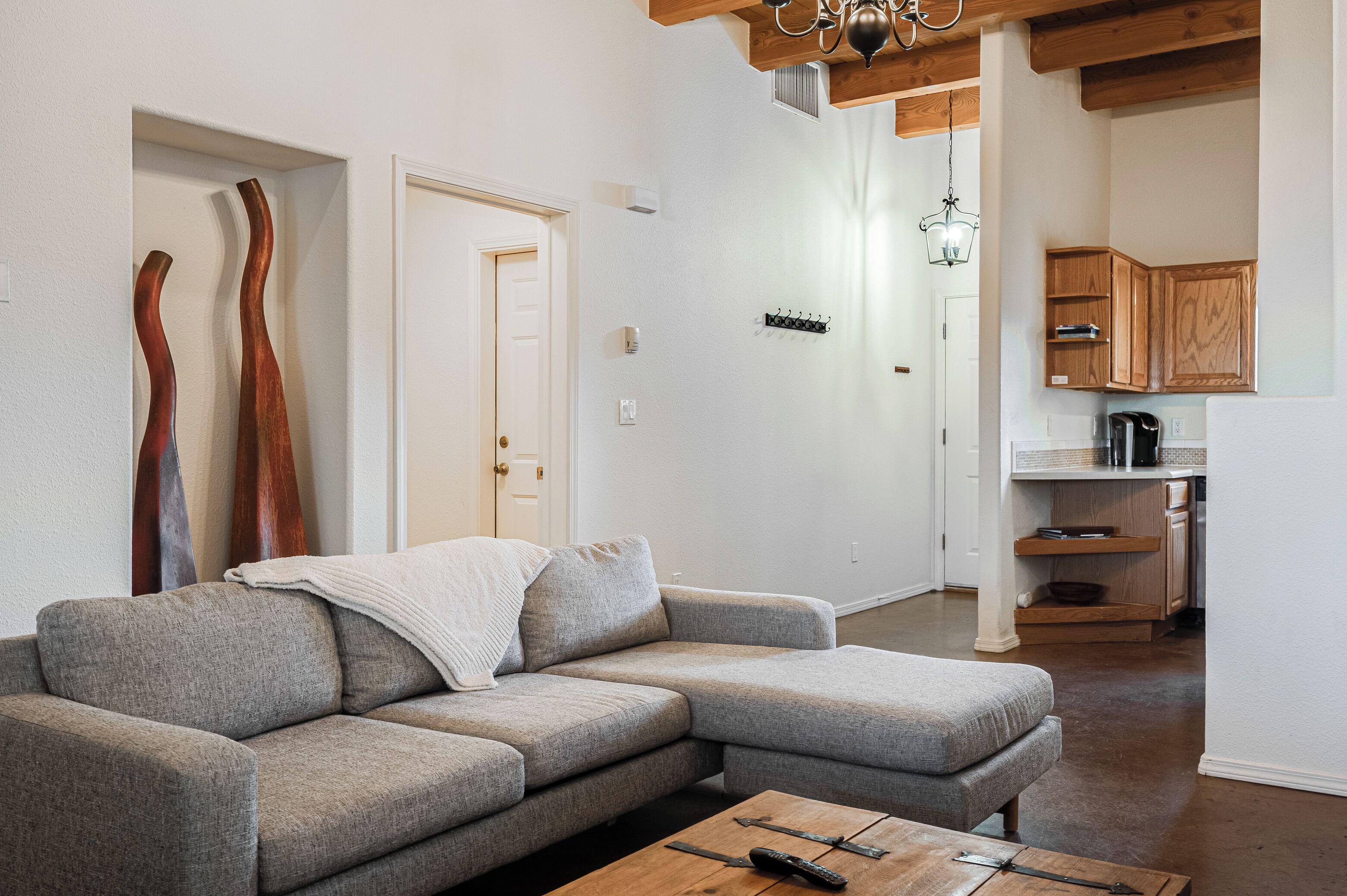 A modern living room with a grey sectional sofa, a wooden coffee table, and a kitchen area with wooden cabinets and a hanging light fixture.