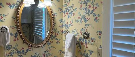 A bathroom with a floral wallpaper, a round mirror with a gold frame, a white sink, and a towel hanging on a rack.