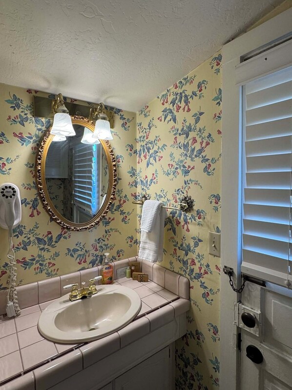 Een badkamer met bloemenbehang, een ronde spiegel met gouden lijst, een wit wastafelmeubel en een handdoek aan een rek.