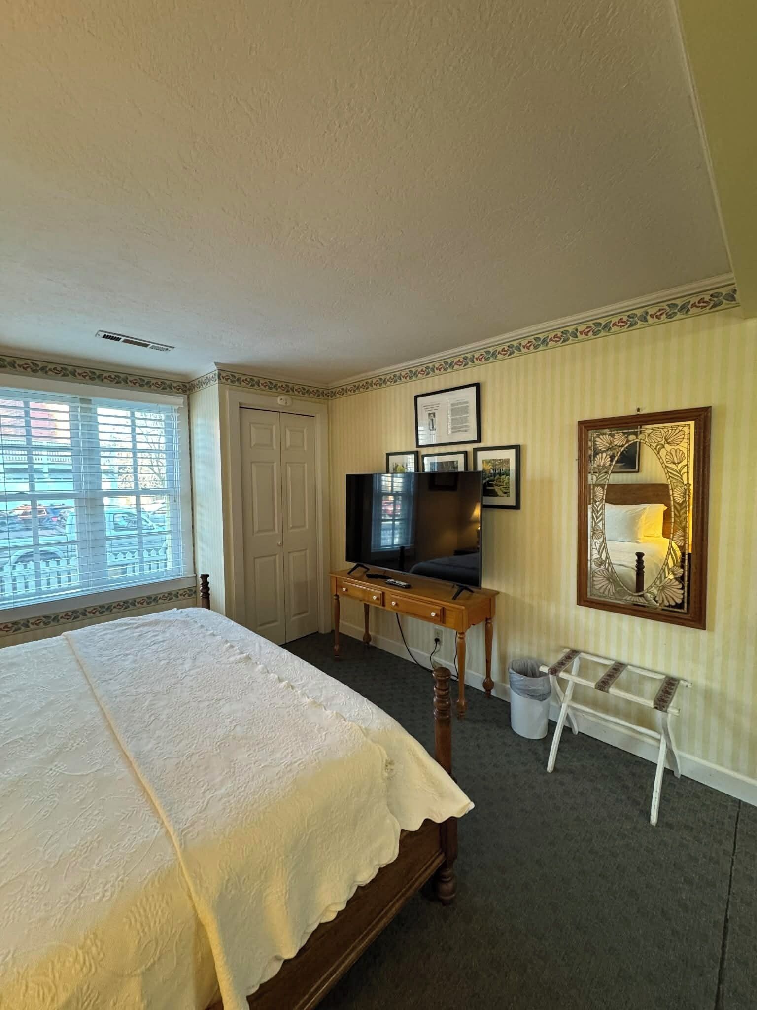 A hotel room with a large bed, a desk with a TV, a mirror, and a window with blinds.