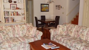 A living room with floral-patterned sofas, a wooden coffee table, a bookshelf, a dining table with chairs, and a chandelier. - Ida s Cottage in Jacksonville (Jacksonville)